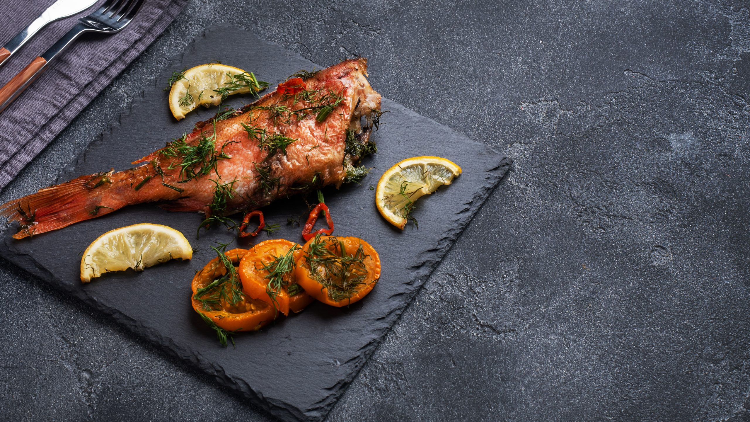 Baked sea bass with tomato pepper lemon and herbs on black slate stand on dark concrete background. Perch fish cooked in the oven Copy space Top view